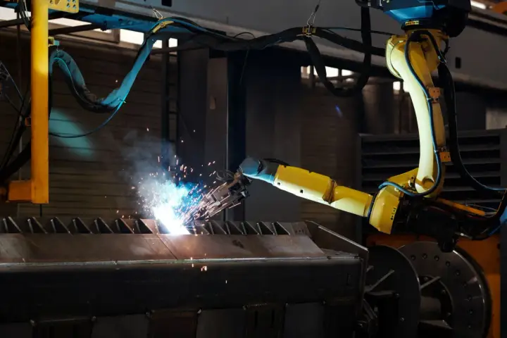 Braccio robotico giallo esegue saldatura, creando scintille luminose su un pezzo metallico grezzo.